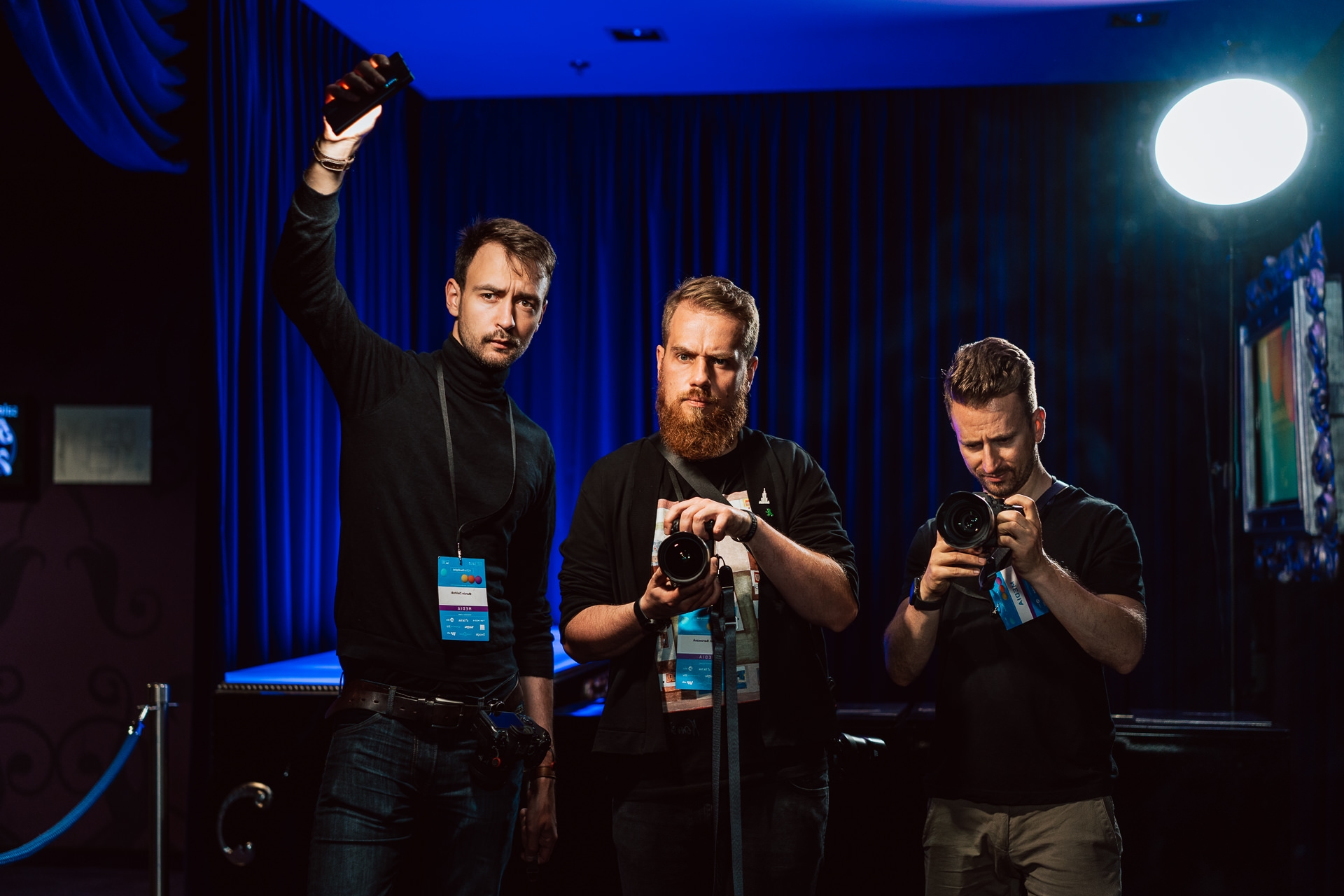 Three men with cameras stand in a room in dramatic lighting; one raises a light source, another photographer at the event holds his camera at chest level, and the third looks down at his camera. In the background are blue curtains and bright studio light.