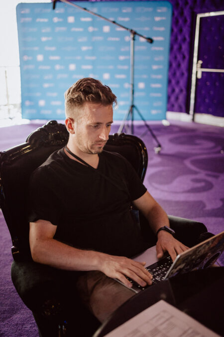 Fotograf na event siedzi w czarnym fotelu, korzystając z laptopa, podczas gdy fotograf na imprezie rejestruje scenę za pomocą niebieskiego banera i statywu mikrofonowego w pokoju z fioletowym dywanem.