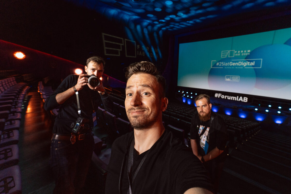 Trzech mężczyzn stoi w pustym audytorium z dużym ekranem wyświetlającym "#ForumIAB" i hashtagi wydarzenia. Fotograf na wydarzeniu robi selfie, inny trzyma aparat, a trzeci stoi za nimi, wszyscy twarzą do kamery.