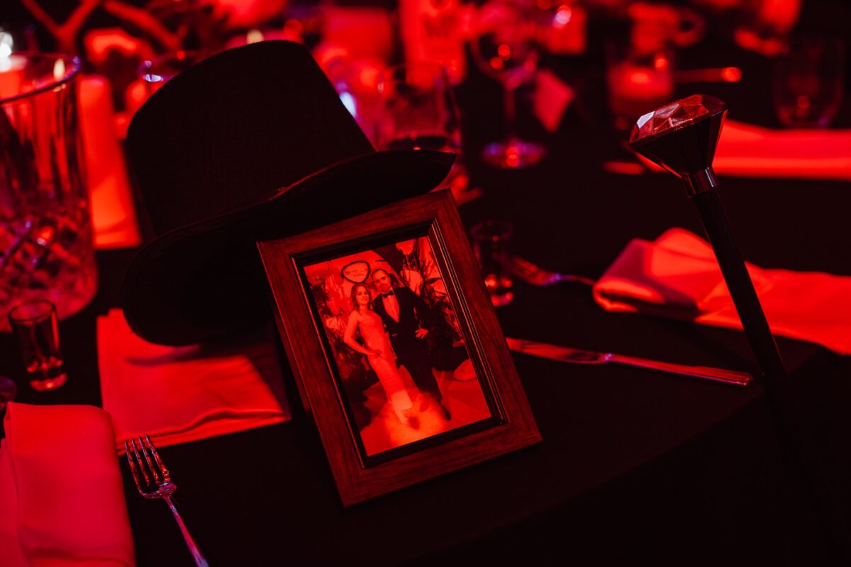 Oprawione zdjęcie wykonane przez fotografa na imprezie przedstawia trzy osoby na stole, obok dużego czarnego nakrycia wierzchniego i laski z rączką w kształcie rombu, wszystko w czerwonym oświetleniu z zastawą stołową i szklankami w tle.