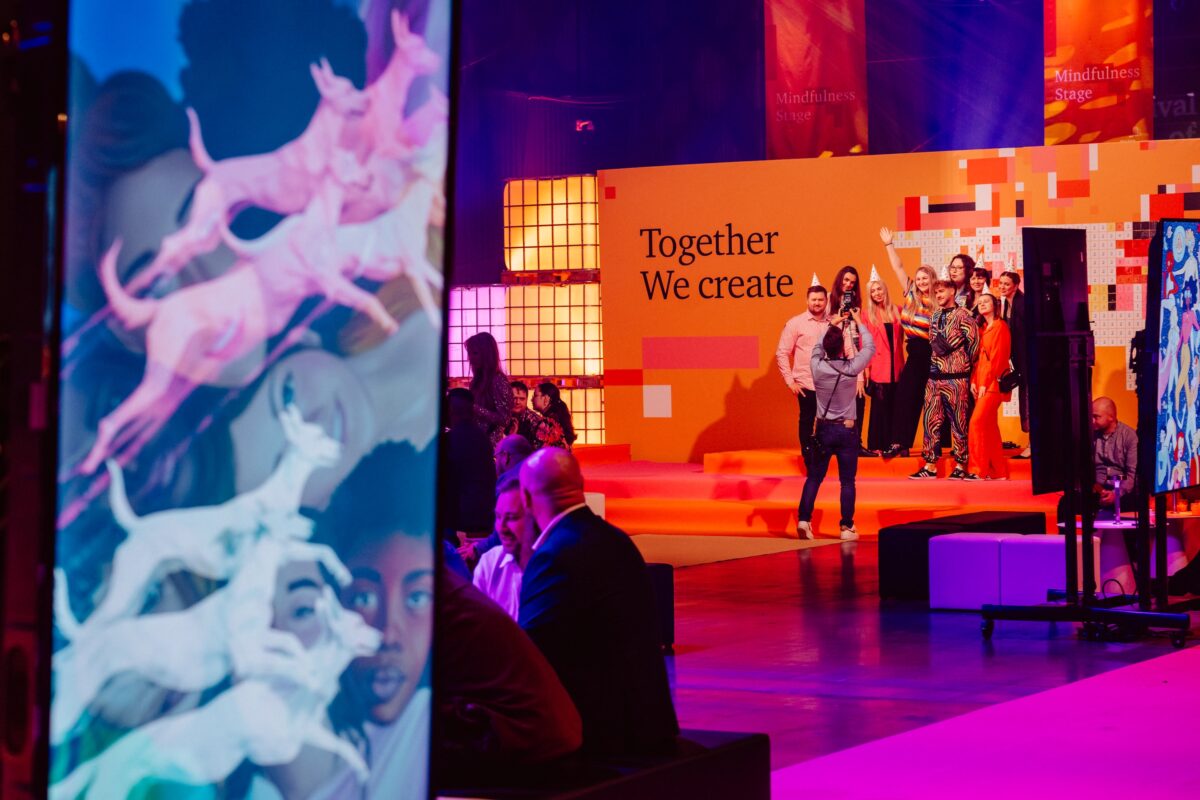 Grupa ludzi stoi na pomarańczowej scenie z napisem "Together We Create" za nimi, podczas gdy inni siedzą i rozmawiają na pierwszym planie. Kolorowa sztuka cyfrowa jest wyświetlana na ekranie po lewej stronie, uchwycona przez fotografa na wydarzeniu.