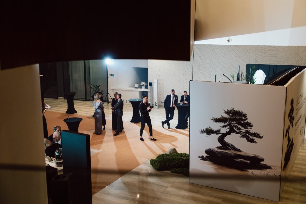 Grupa dobrze ubranych osób przechodzi przez nowoczesne, eleganckie lobby z dużym obrazem drzewka bonsai. Fotograf na imprezie rejestruje scenę, gdy mężczyzna w garniturze przemawia, a inni obserwują, otoczeni wypolerowanymi podłogami i oświetleniem otoczenia.