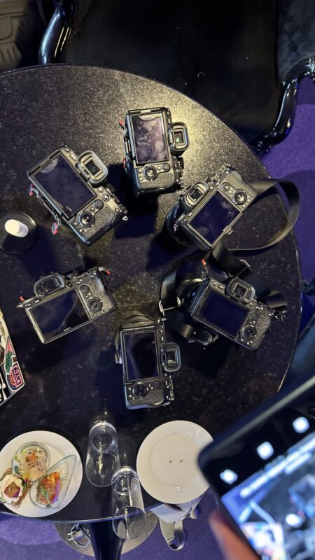 Sześć aparatów cyfrowych z ekranami LCD skierowanymi do góry ustawionych jest w okręgu na okrągłym czarnym stole, co sugeruje, że jest to impreza fotograficzna. W pobliżu znajdują się puste talerze, szklanki, trochę jedzenia i część smartfona na krawędzi obrazu.