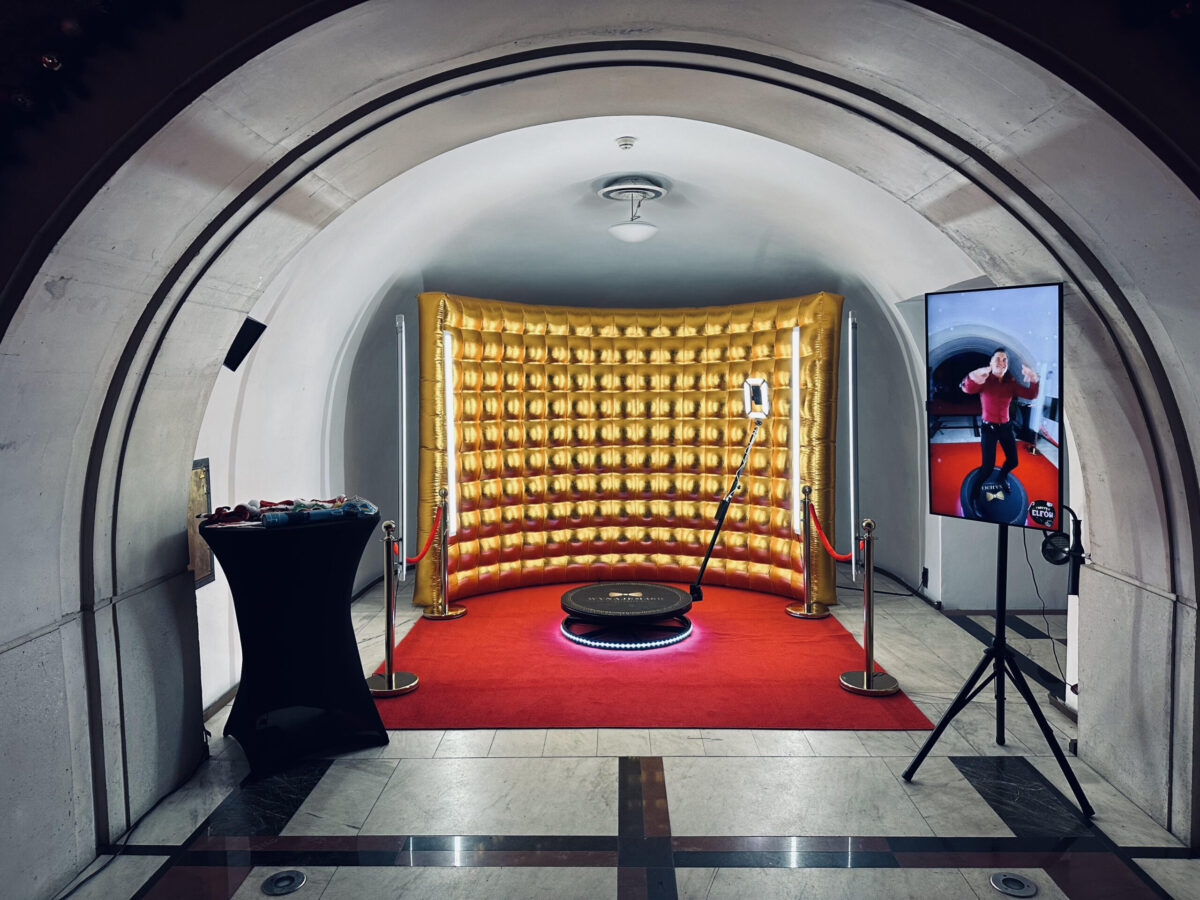 Galeria 6 scaled 1 Fotobudka ze złotym nadmuchiwanym tłem, czerwonym dywanem i kłonicami pod łukowym sufitem. Z przodu znajduje się czarna okrągła platforma i aparat na statywie, a na wysokim ekranie wyświetlana jest pozująca osoba.