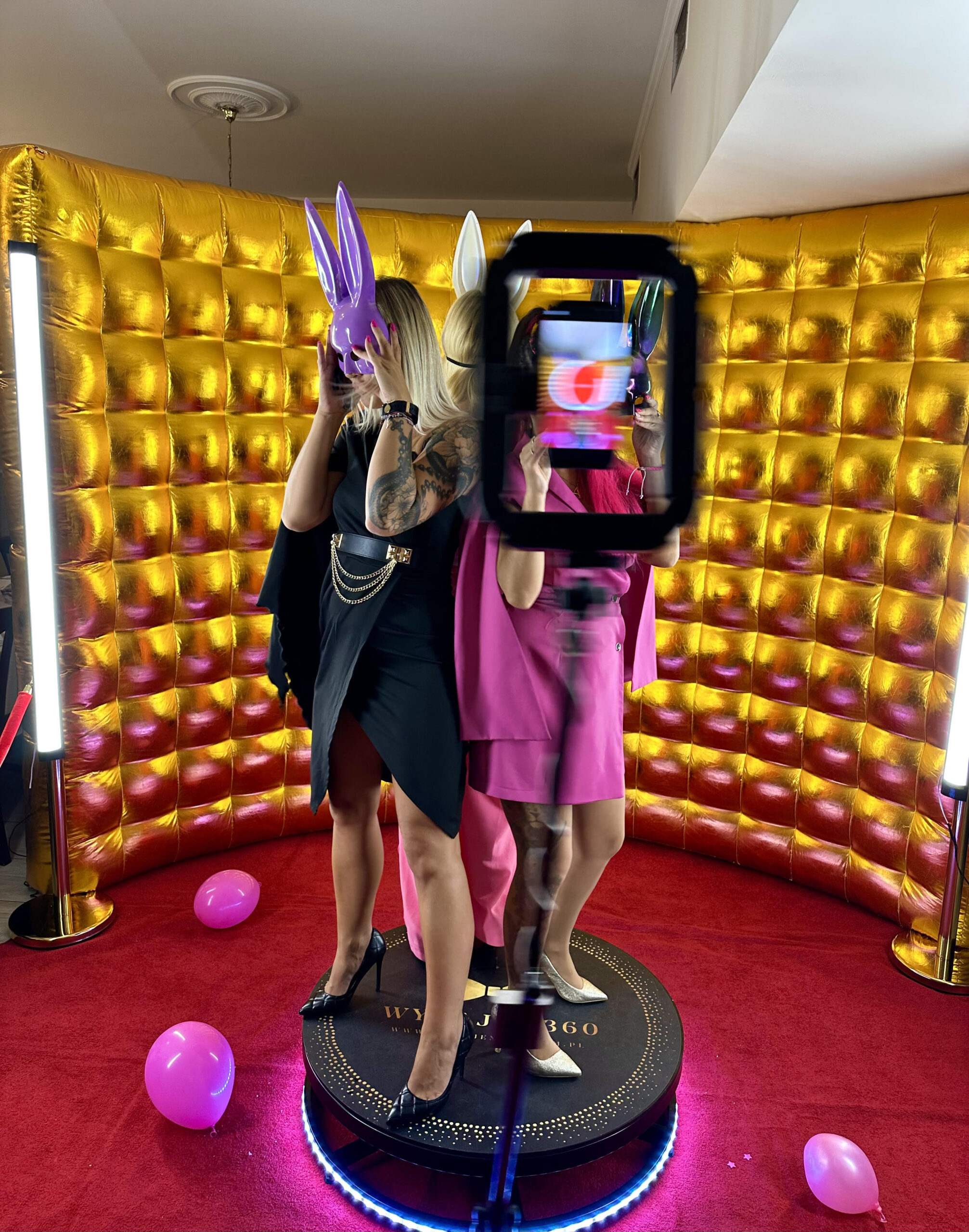 IMG 8242 scaled 1 Wynajem Fotobudki 360 - Budka fotograficzna z okrągłą platformą i światłem pierścieniowym stoi na czerwonym dywanie przed złotym, pikowanym tłem, otoczonym dwoma pionowymi światłami LED w słabo oświetlonym pomieszczeniu.