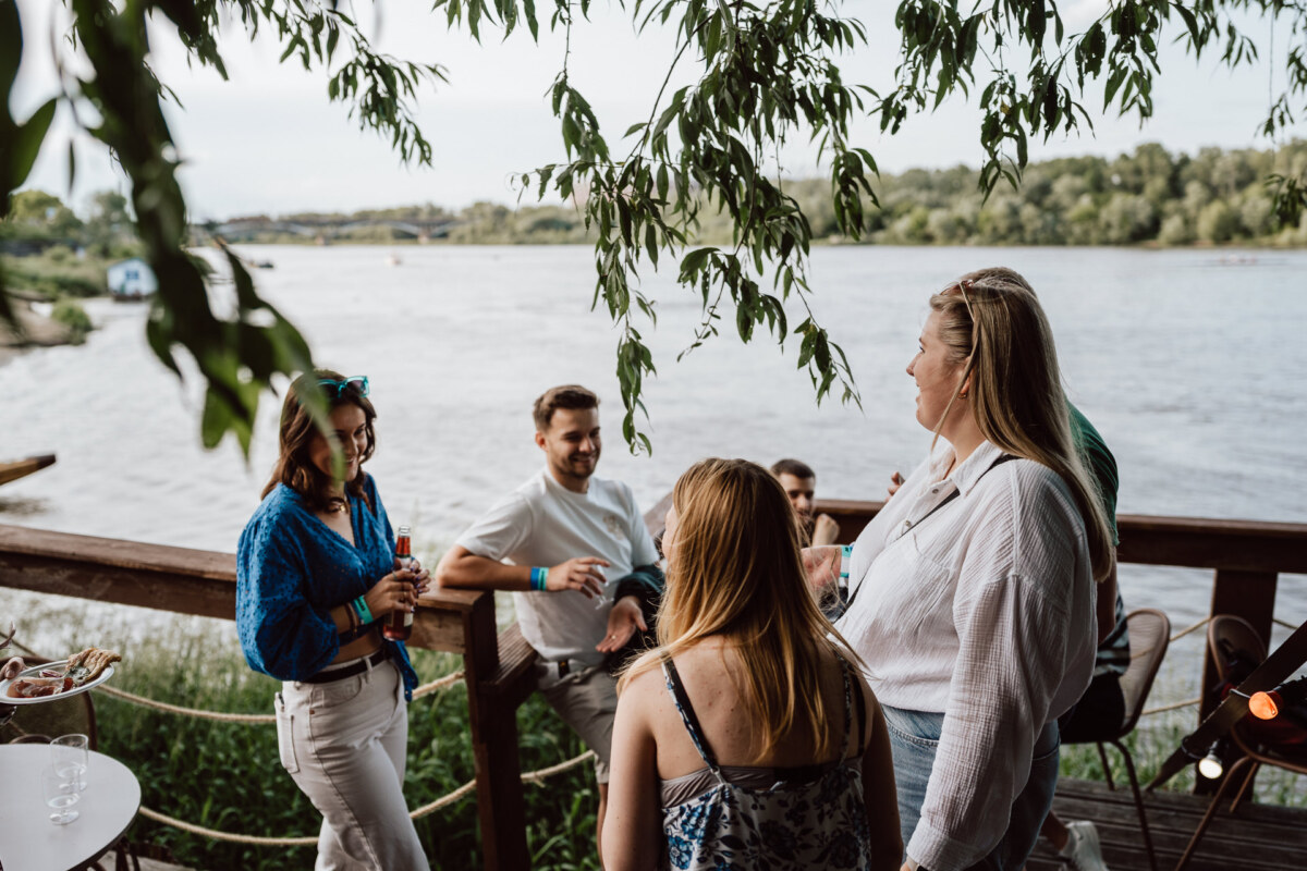 A group of people are meeting outdoors on a wooden terrace overlooking the river, with trees in the background and leafy branches hanging above. Some are holding drinks and engaged in conversation. 