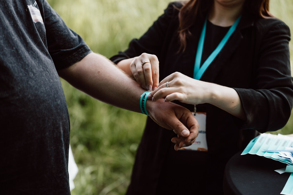 Outside, a person puts a turquoise-colored wristband on the wrist of another person. Both people are standing at a table, and one of them is wearing a lanyard with an ID badge. The background is a fuzzy green.  