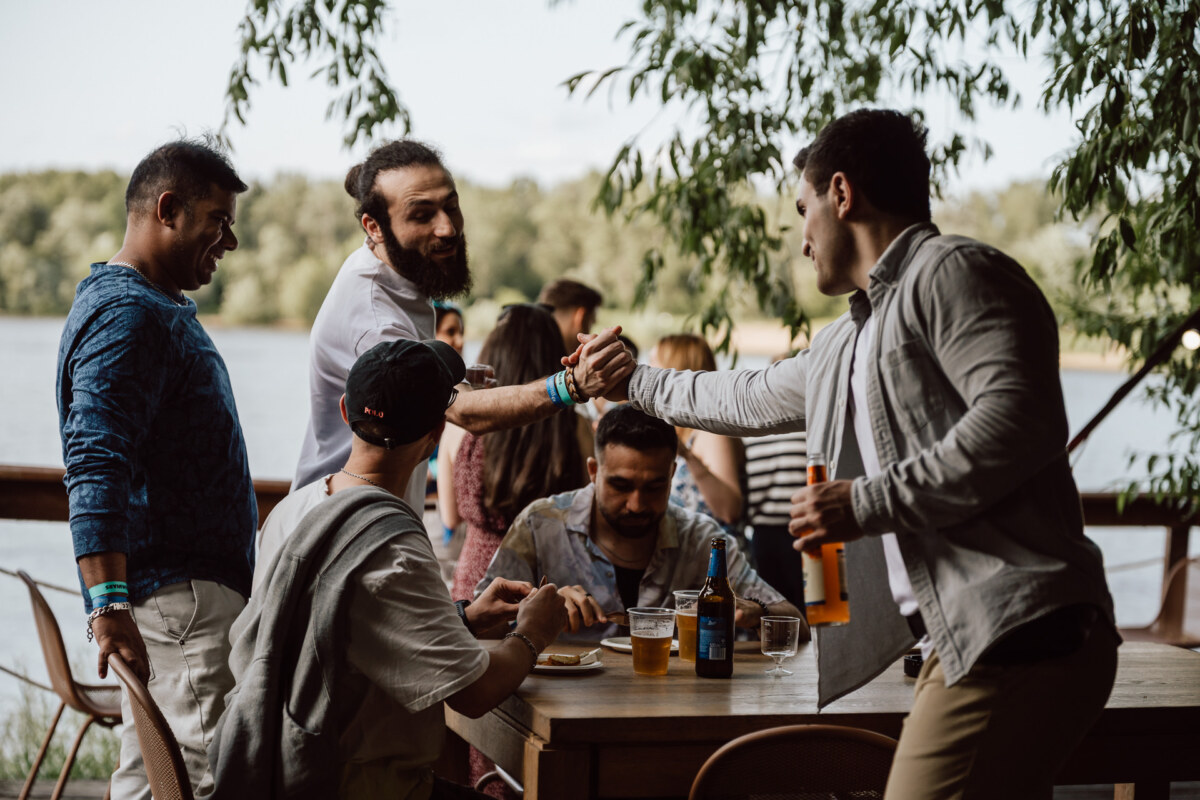 A group of friends gather around an outdoor table by the lake, smiling and shaking hands. Some sit with drinks and food, while others stand and engage in friendly conversation surrounded by trees. 