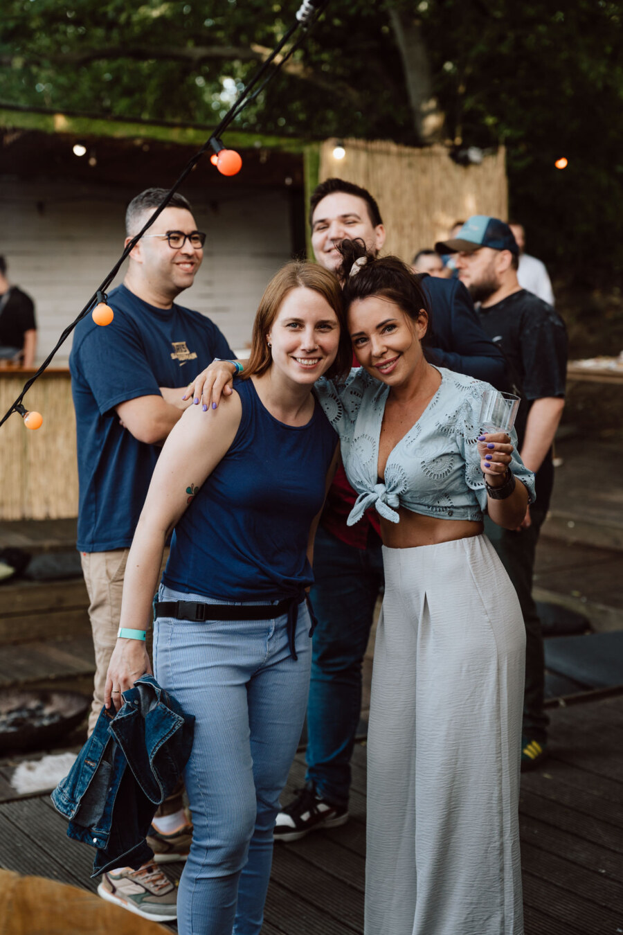 Two women pose together and smile during an outdoor gathering, one of them holding a drink. Several people stand and talk in the background under string lights, with trees and a wooden structure visible. 