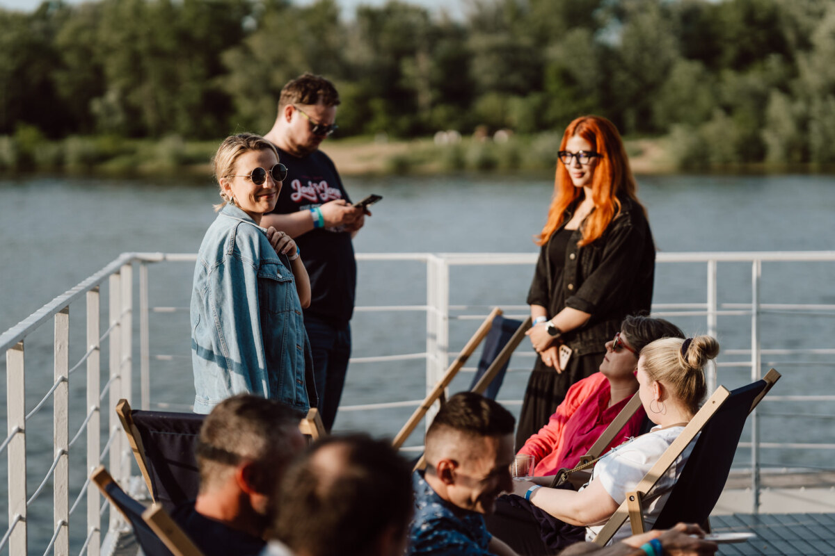 A group of people are relaxing on deck chairs on a boat, with trees and water in the background. Some are talking, one person is looking at his phone, and another is smiling at the camera wearing sunglasses. 