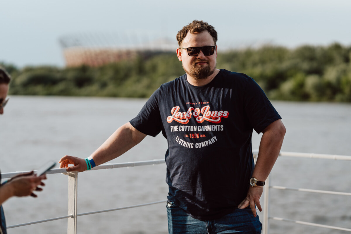 A man wearing sunglasses and a "Jack &amp; Jones" T-shirt is standing by a railing over a river, with trees and a stadium visible in the blurry background. To the left, the hand of another person can be seen holding a phone. 
