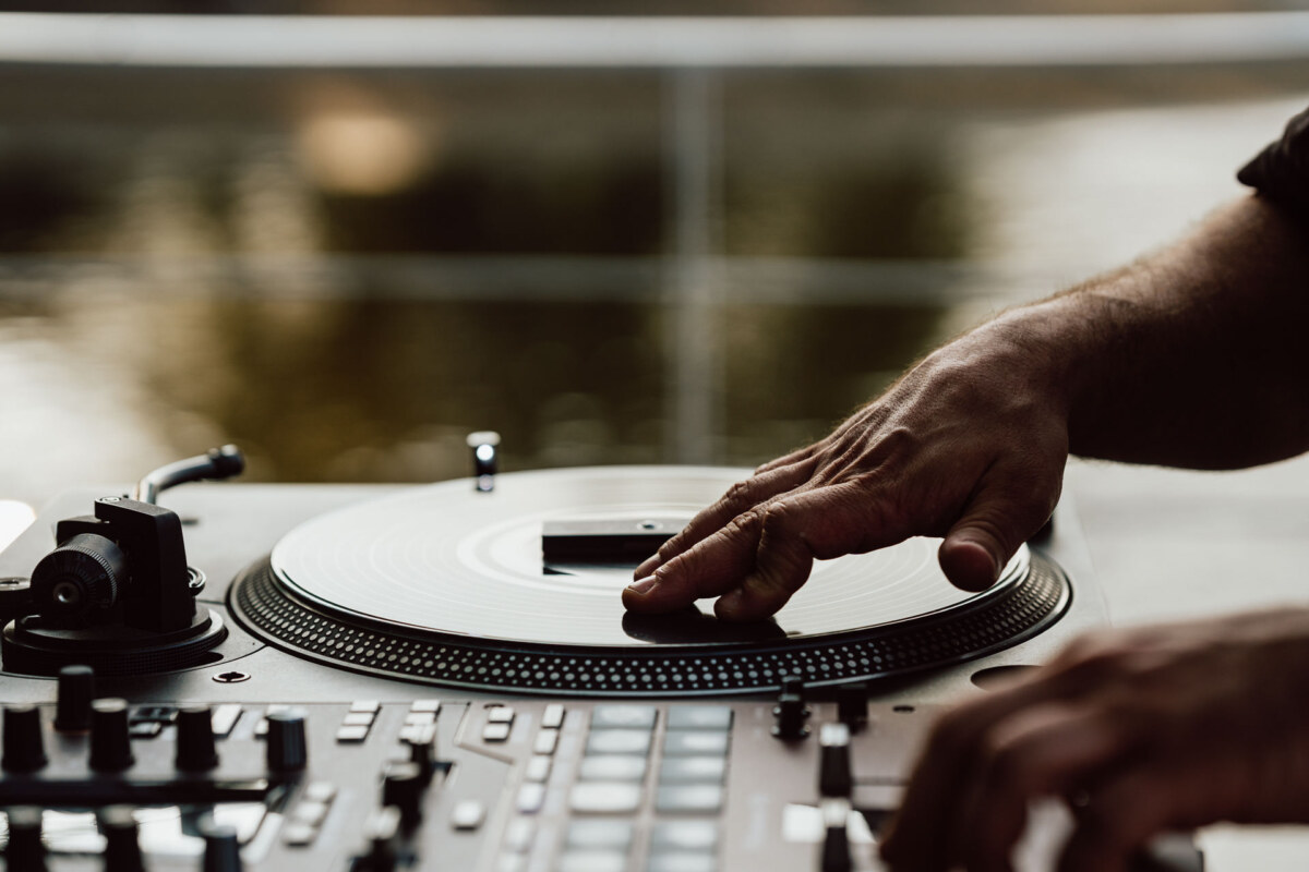 Close-up of the hands of the person operating the turntable and DJ mixer, with one hand touching the vinyl record and the other adjusting the controls. The background is blurred, suggesting an outdoor setting. 