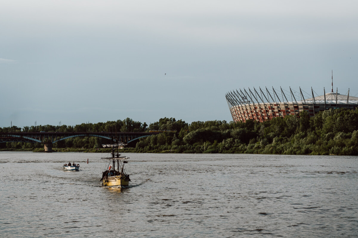A yellow boat and a smaller white boat sail on the river near the bridge, with a large stadium and green trees under an overcast sky visible in the background.