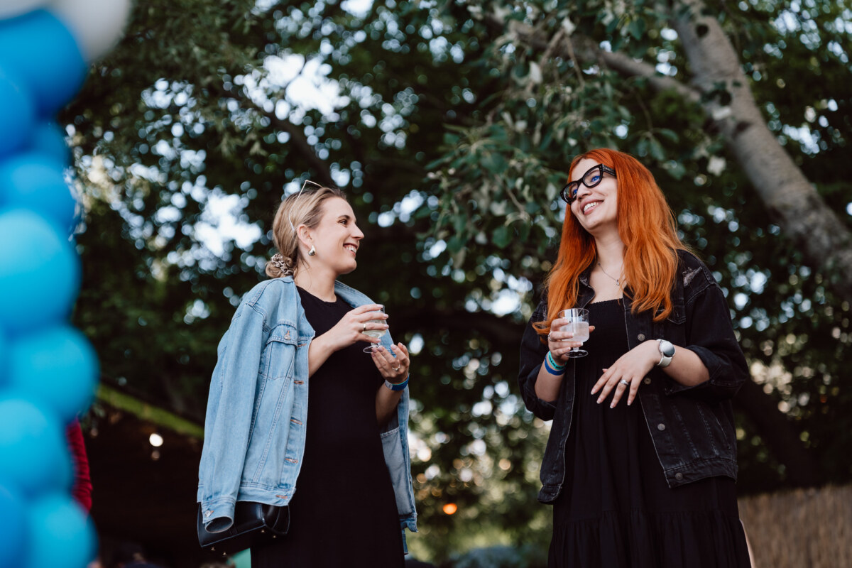 Two women are standing outside, smiling and talking, holding drinks. One has blond hair and wears a denim jacket; the other has long red hair, glasses and a black jacket. Trees and blue balloons are visible in the background.  