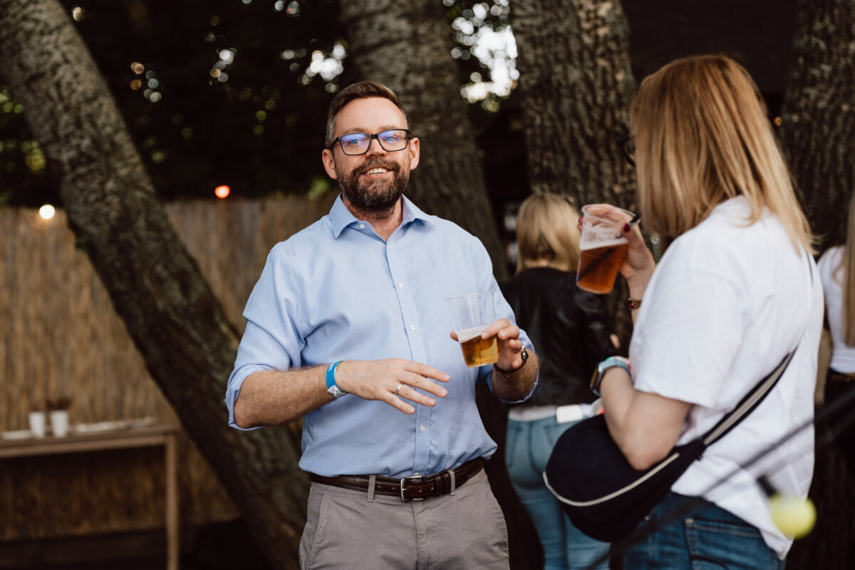 A man in glasses and a blue shirt smiles while holding a drink and talking to a woman holding a beer at an outdoor meeting, with trees and a wooden fence in the background.