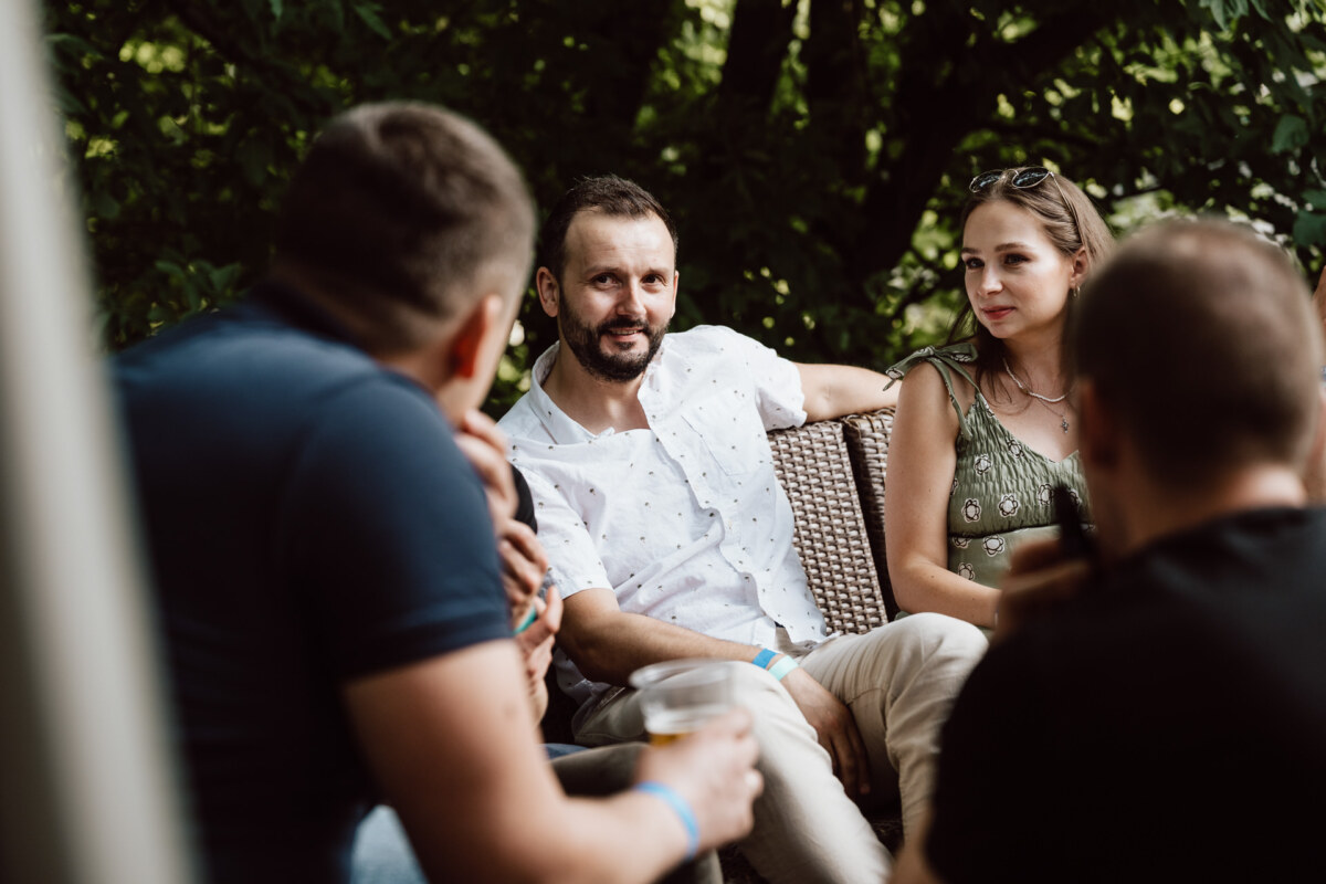 A group of people sit close together outside, engaged in conversation. One man in a white shirt speaks while the others listen. The setting is casual, with trees and greenery in the background.  