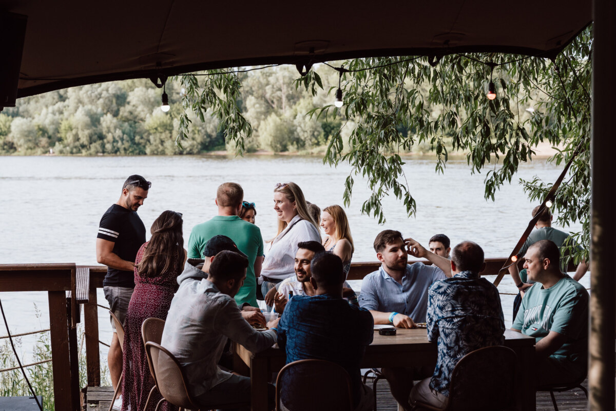 A group of people gather and talk at tables on a shaded terrace by the river, with trees and greenery in the background on a sunny day.