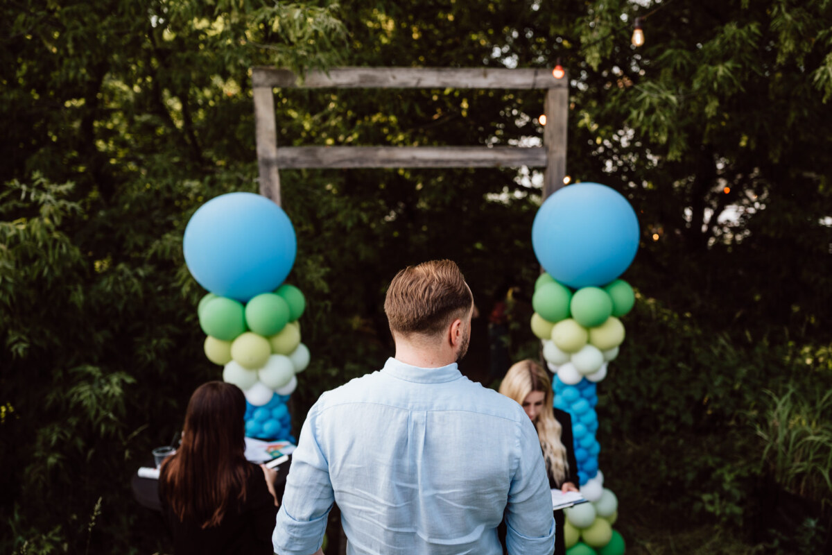 A man in a light blue shirt stands outside in front of a wooden arch decorated with blue, green and white balloons, while two people surrounded by greenery stand in front of him.