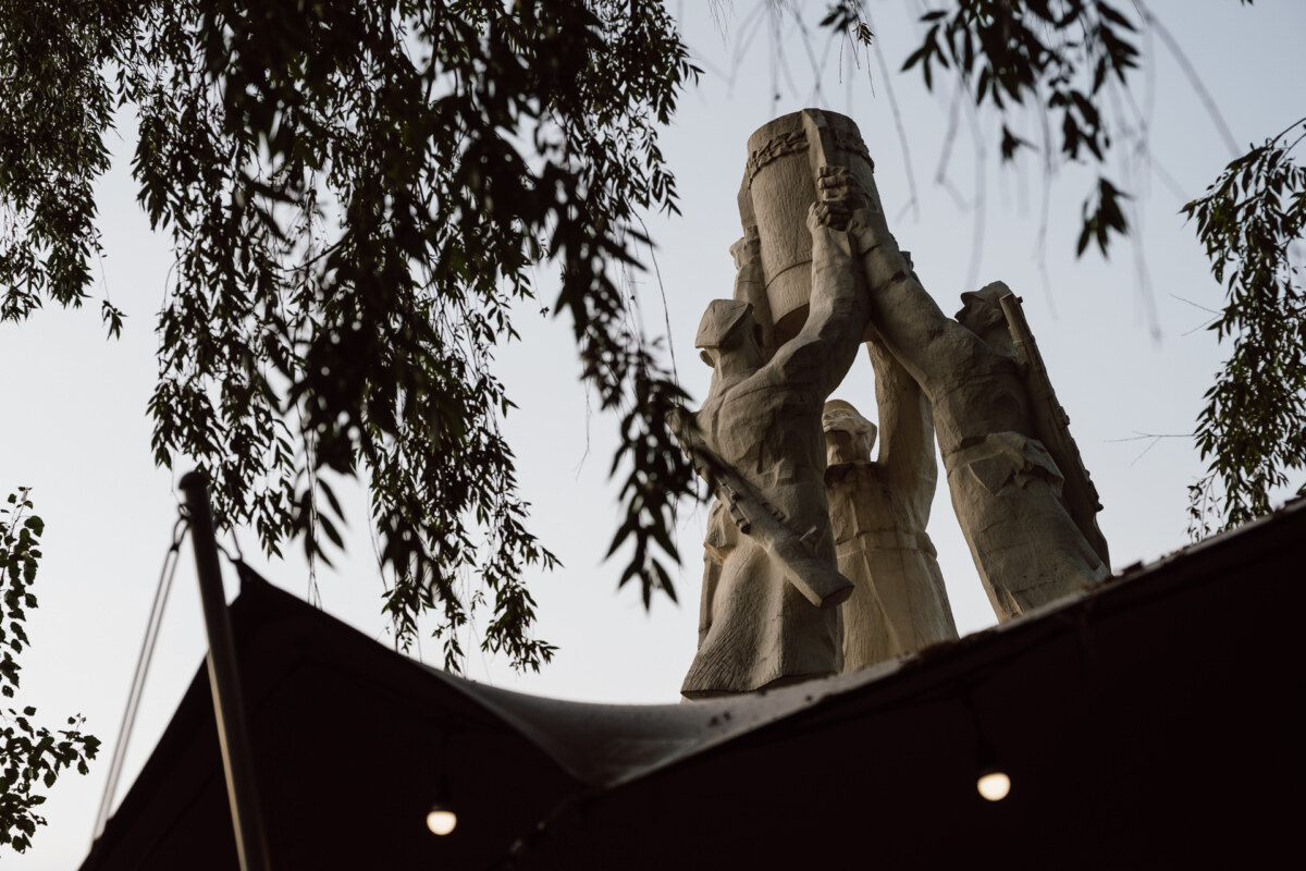 A stone monument depicting four figures lifting a large object together, partially framed by leafy tree branches and seen from a low angle against the sky.