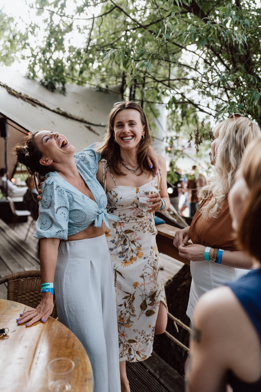 Four women are standing together outside, smiling and laughing next to a wooden table under the trees. They appear to be enjoying a casual get-together, dressed in light summer clothes and holding drinks. 