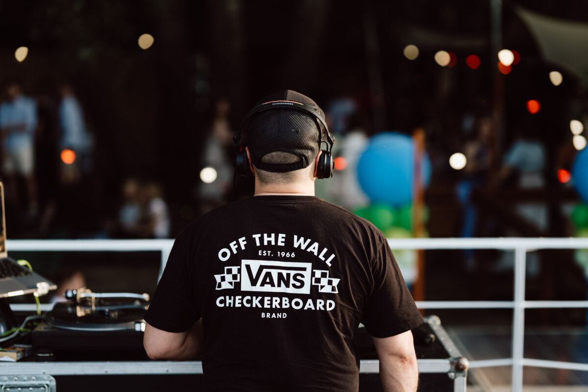 A DJ wearing headphones and a black "Vans Off the Wall" T-shirt stands at a turntable, performing outdoors with fuzzy people and colorful balloons in the background.