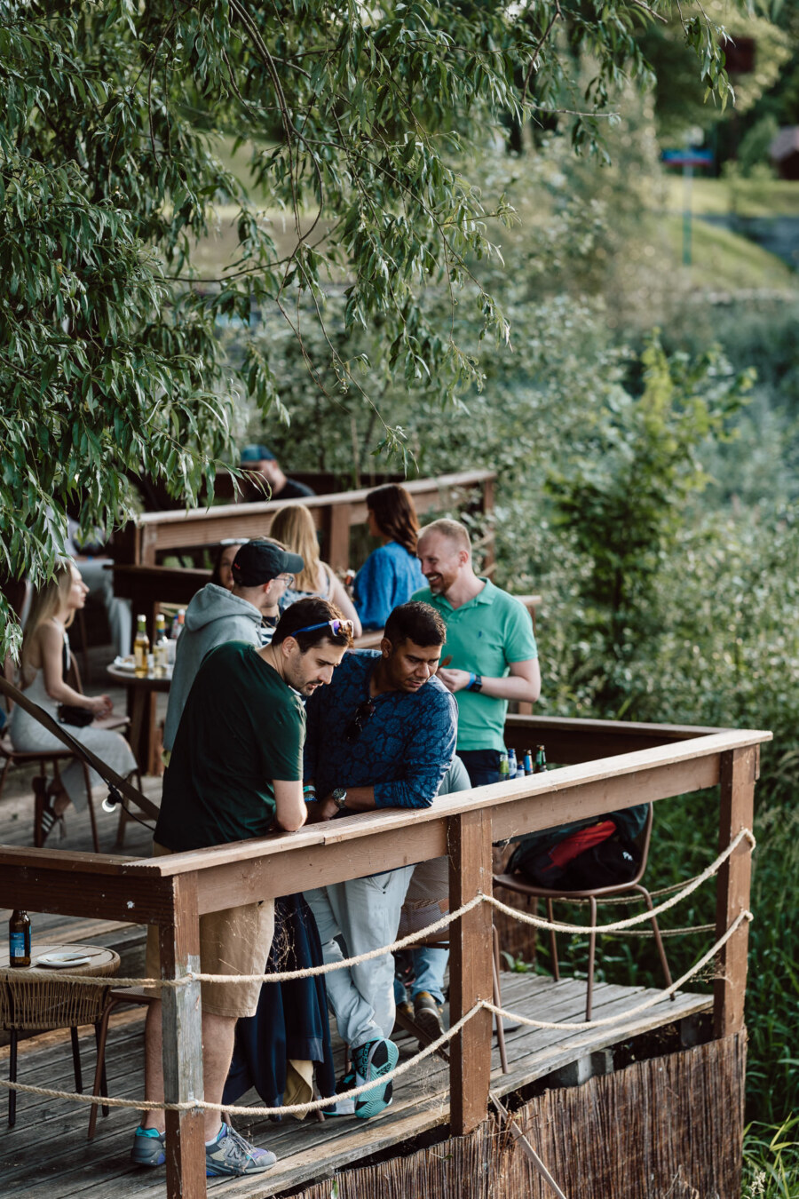 A group of people on a wooden terrace surrounded by green trees and foliage, with drinks and snacks on tables. Some stand and talk, while others sit, enjoying the casual atmosphere. 