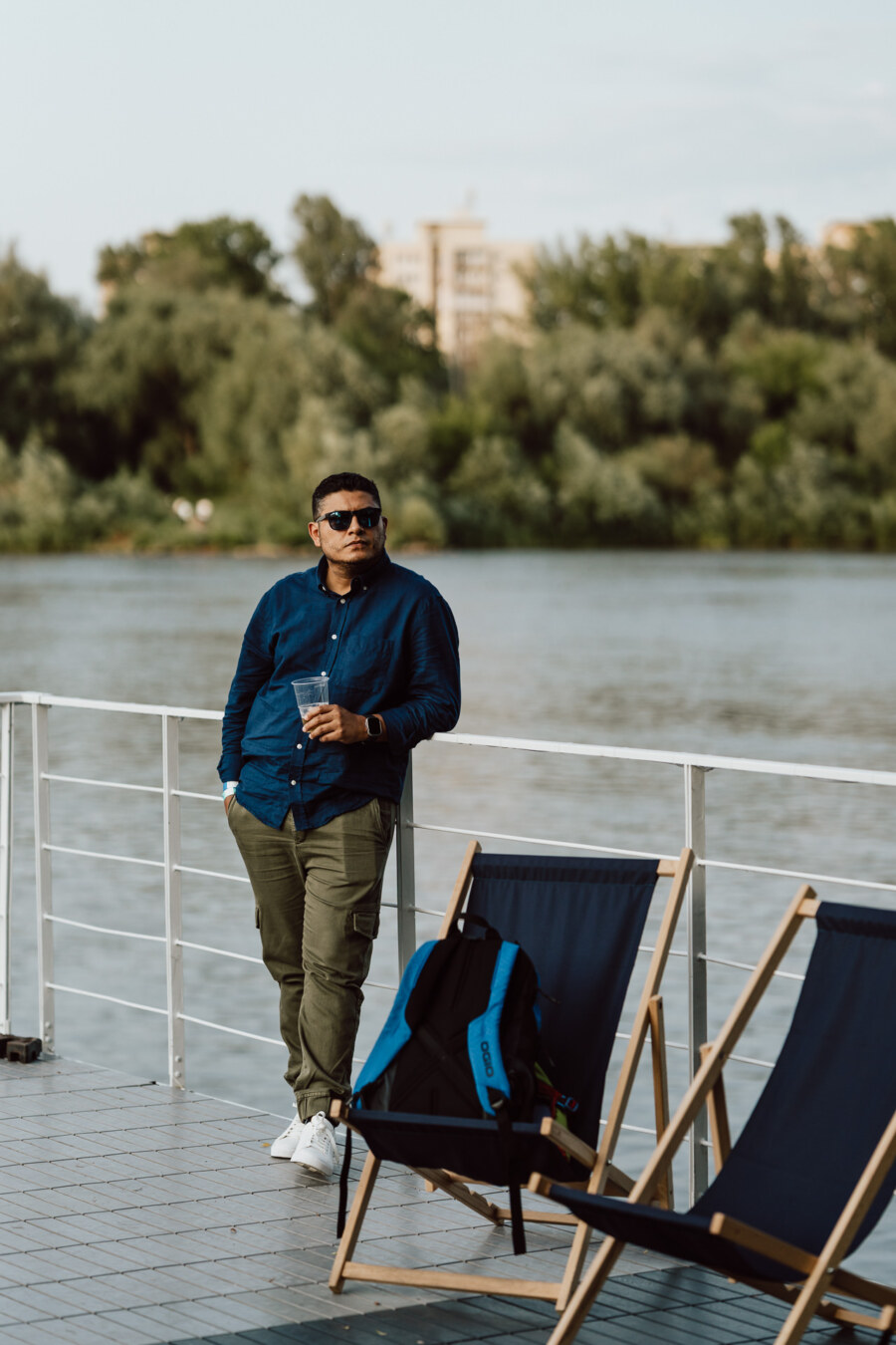 A man wearing sunglasses and casual clothes leans against the railing by the river, holding a drink. Nearby are two empty deck chairs and a backpack. Trees and buildings are visible in the background.  