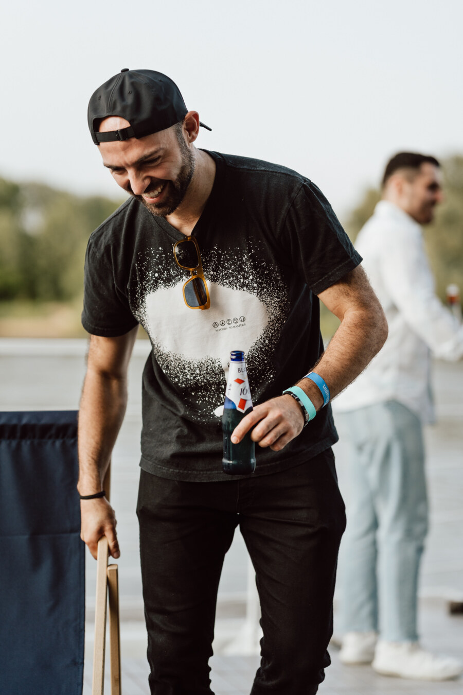 A bearded man wearing a black cap and T-shirt smiles while holding a beer bottle out. He wears sunglasses on his shirt and has a wrist band. Another person stands vaguely in the background.  