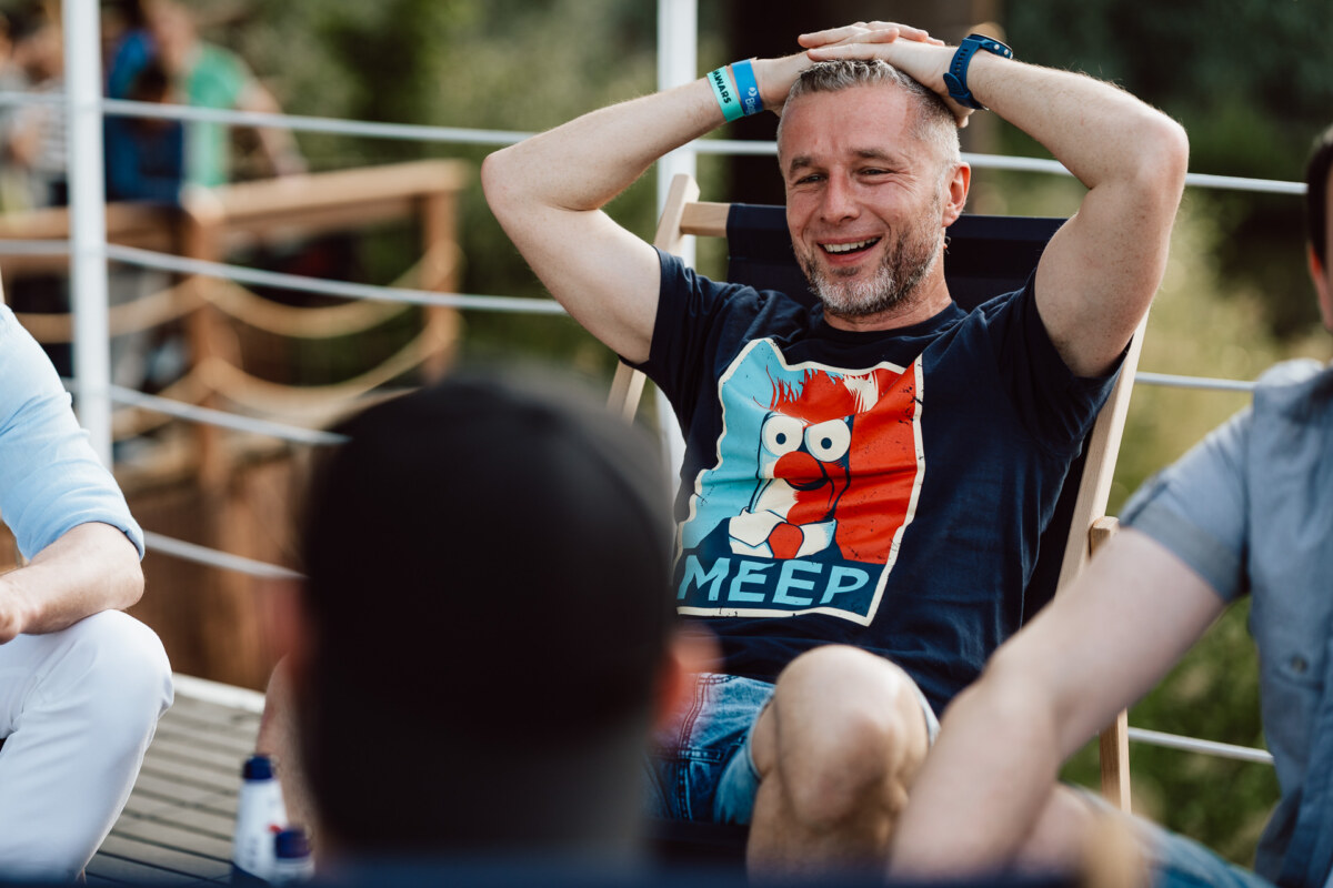 The man is sitting outside on a deck chair, smiling with his hands resting on his head. He is wearing a dark T-shirt with a cartoon character and "MEEP" written on it. Two other people are partially visible in the foreground.  
