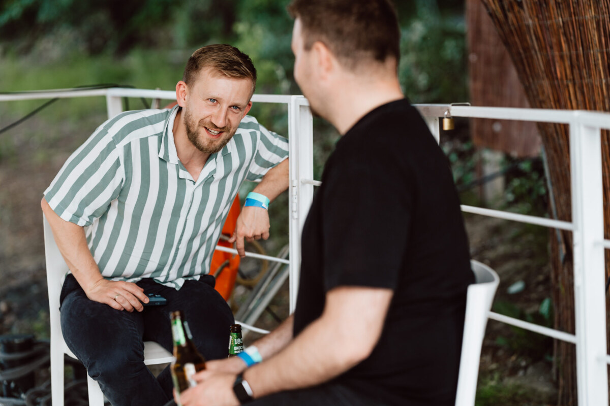 Two men sit and talk outside; one, wearing a green and white striped shirt, smiles, leaning forward and holding a drink. The other, wearing a black shirt, sits facing away, also holding a drink. The green is visible in the background.  