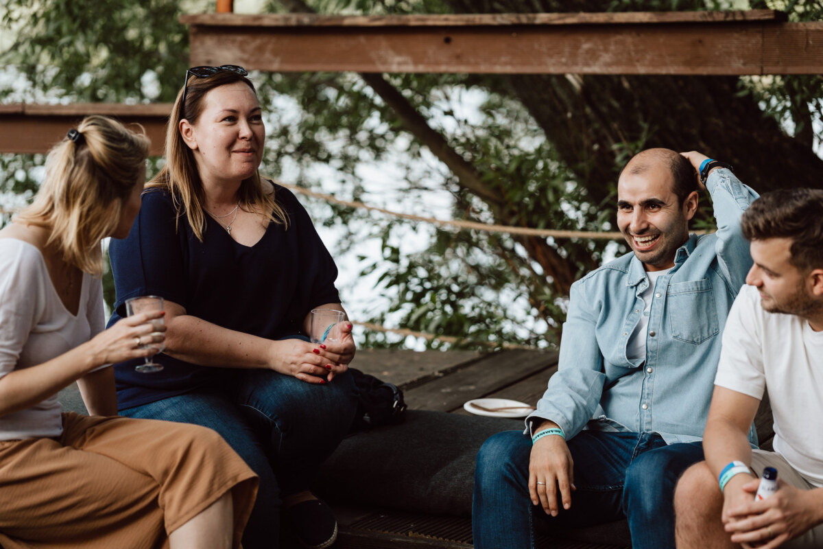 Four adults are sitting and talking outdoors on a wooden terrace near the trees. One person is holding a drink, and everyone seems relaxed and engaged in conversation. 