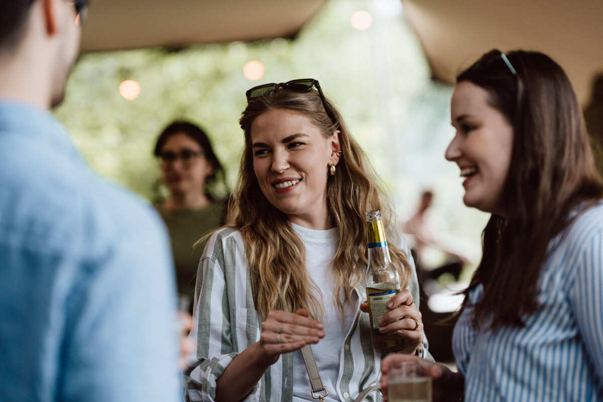 Three people are standing together outside, smiling and talking. One of the women in the center is holding a bottle of drink. Two women and one man are visible, with blurred people and trees in the background.  