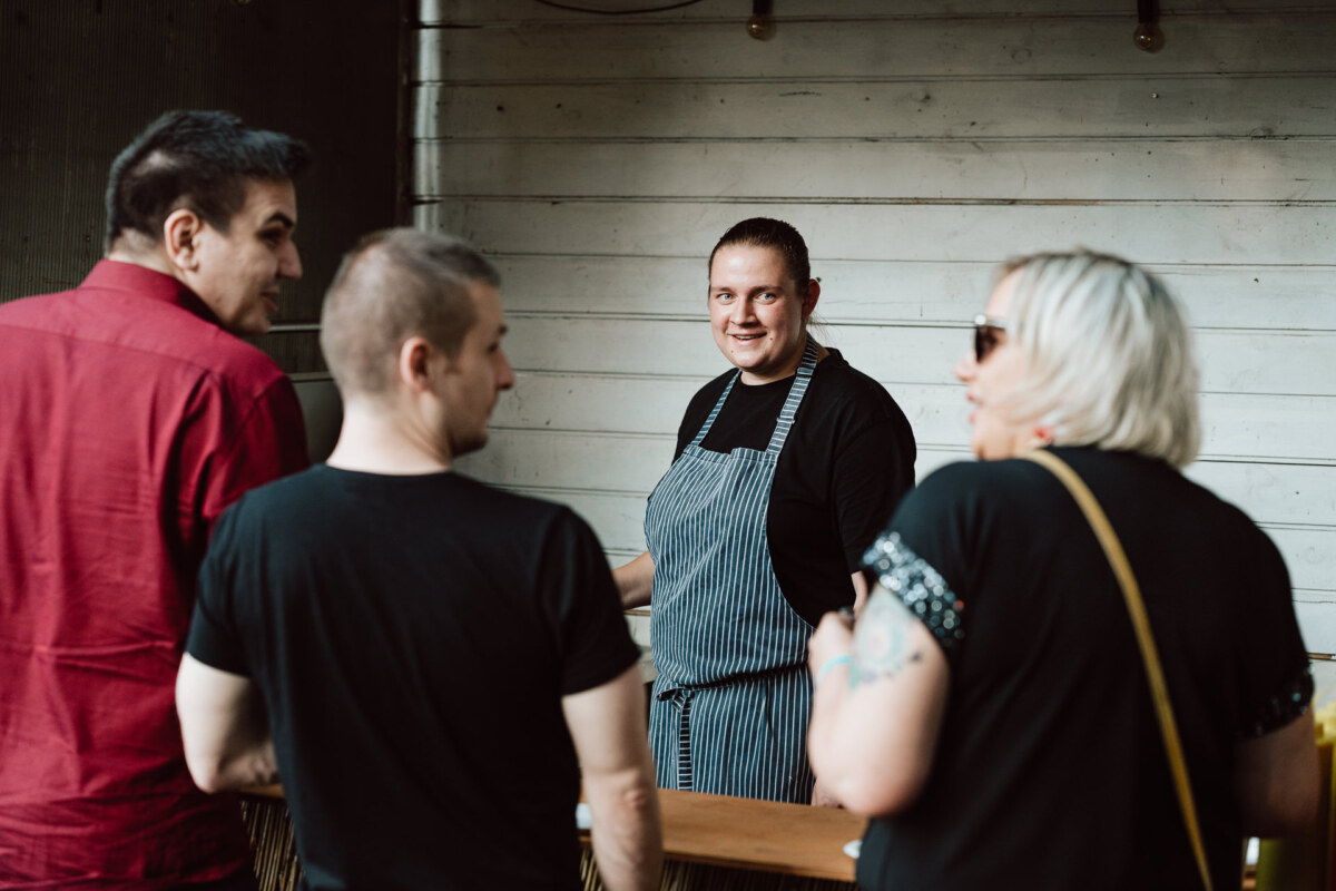 A chef in a striped apron smiles at three people standing at the counter outside, with a white wooden wall in the background.