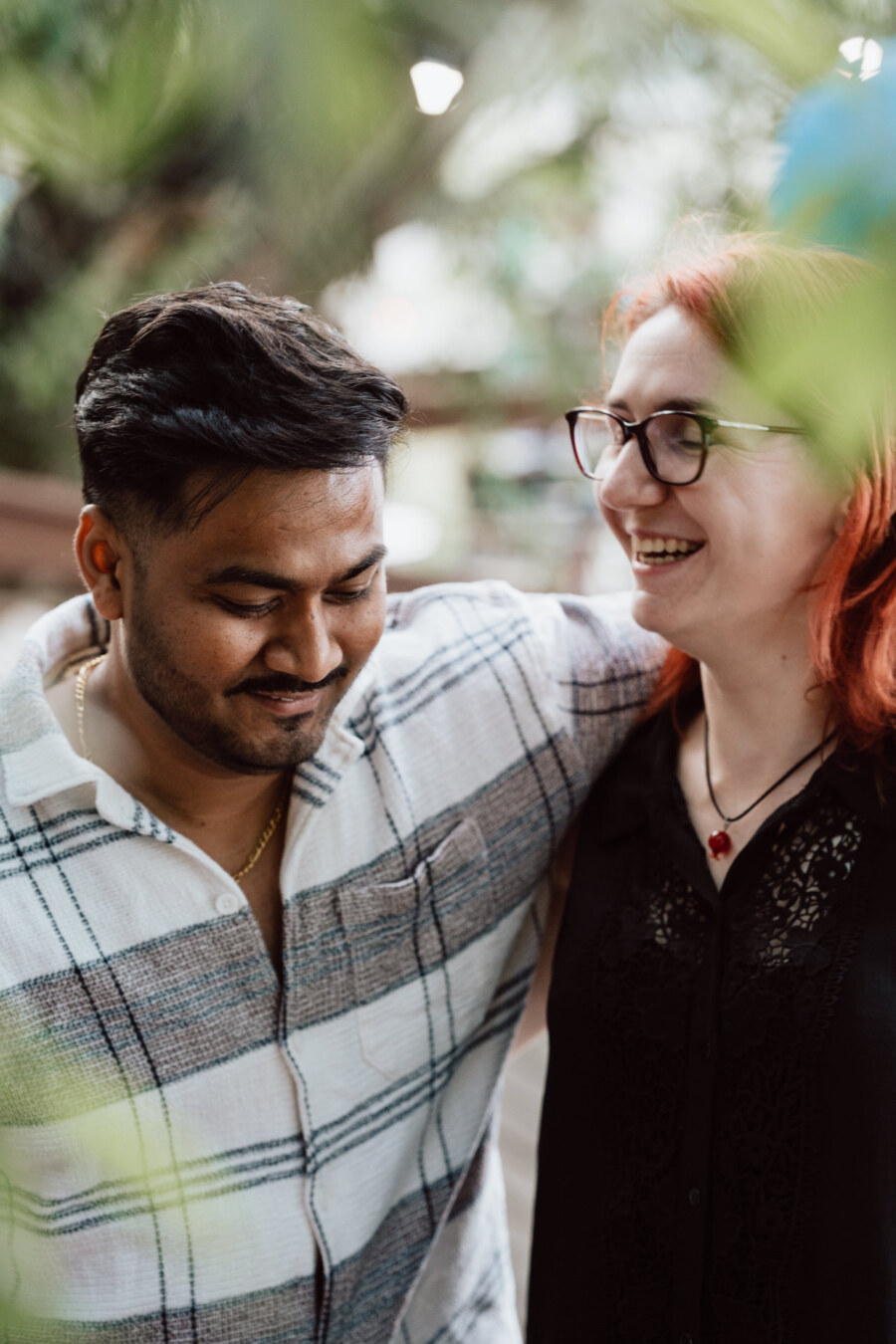 A man and a woman stand close together outside, smiling and laughing. The woman puts her arm around the man's shoulder; both seem happy and relaxed, surrounded by greenery. 