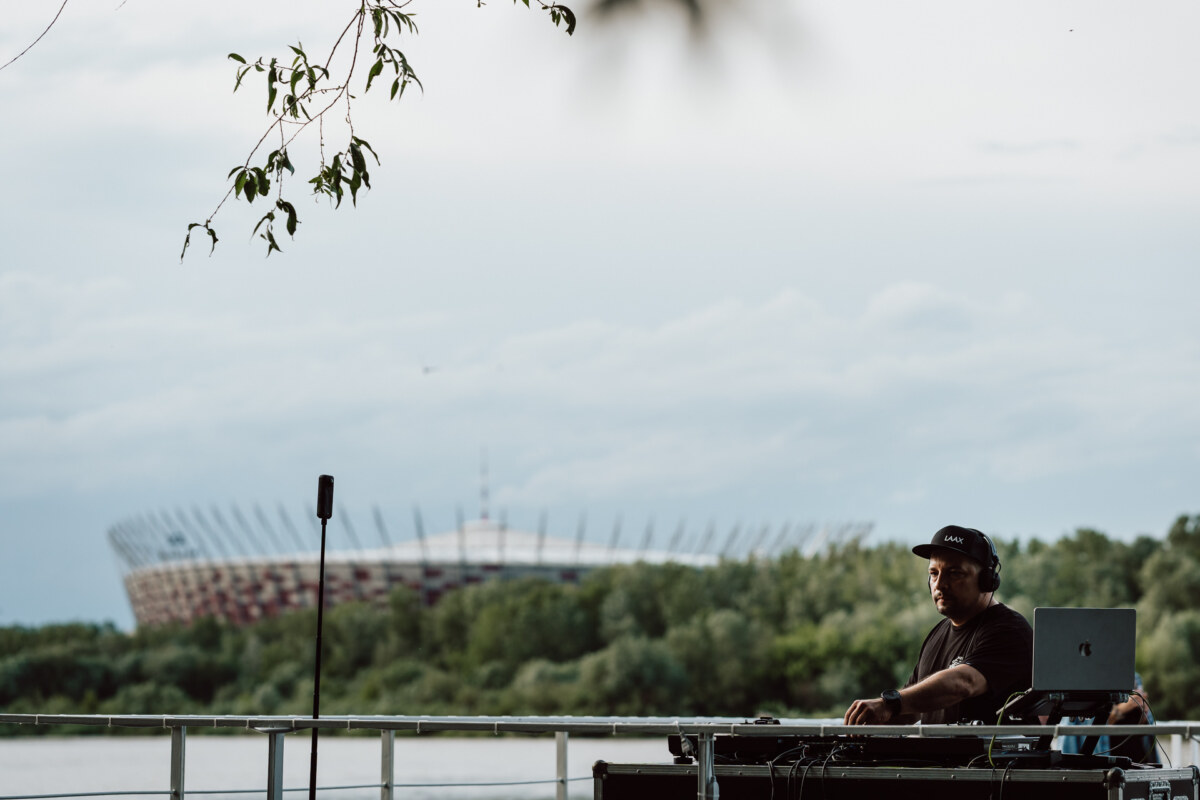 The DJ, wearing a black shirt and cap, performs on outdoor decks by the river, with trees and a large stadium visible in the background under an overcast sky.