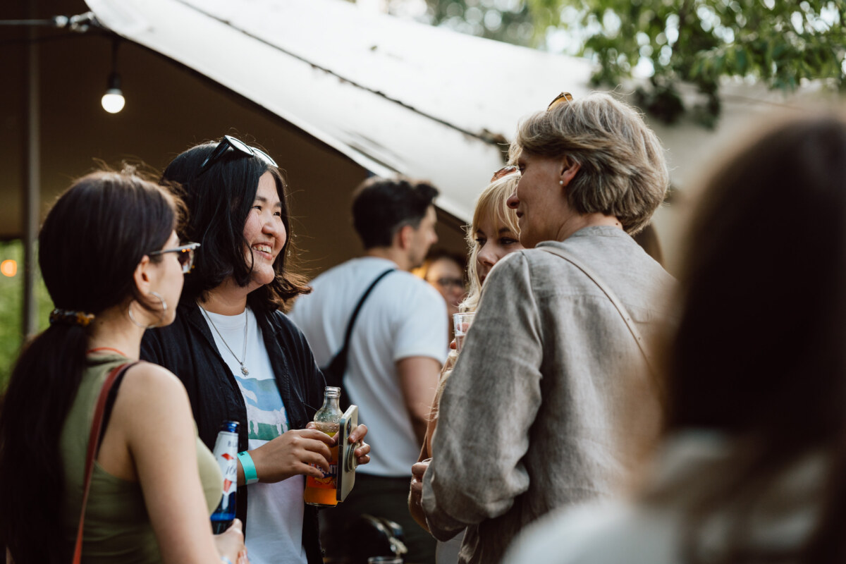 A group of women stand outside under a tent, talking and smiling, holding drinks. Trees and other people are visible in the background, suggesting a relaxed social gathering or event. 