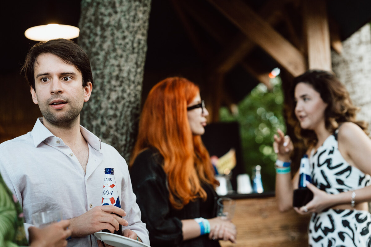 A man in a white shirt holding a drink stands outside near two women, one with long red hair and glasses, both engaged in conversation. The setting appears to be a casual social gathering. 
