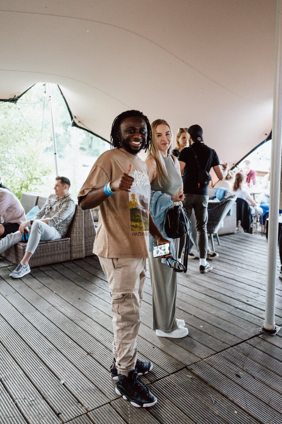 The man smiles and lifts his thumb up, standing next to the woman, who is also smiling. They are at an outdoor event under a tent with people sitting and socializing in the background. 