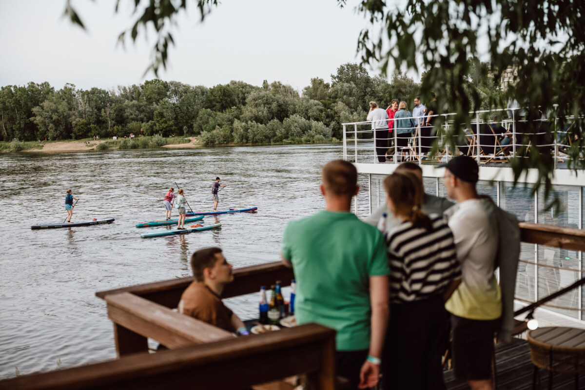 People watch from a riverside terrace as several paddleboarders glide across the wide river, surrounded by trees. Some sit and others stand, enjoying the view on a sunny day. 