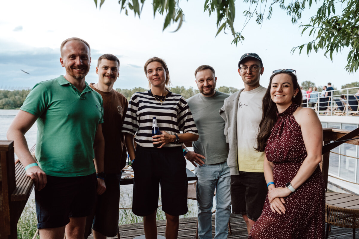 Six people stand together and smile for a group shots outdoors by a lake, with trees, water and a wooden pier visible in the background.