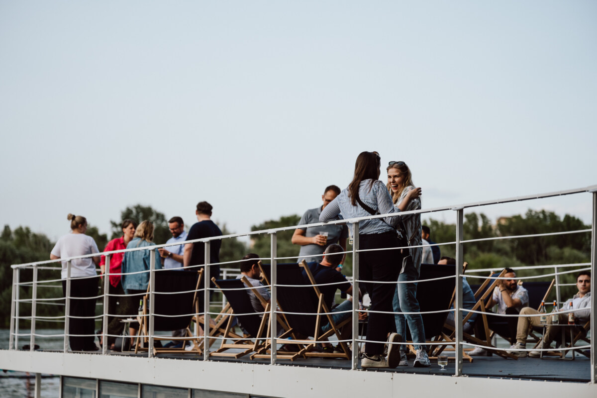 A group of people meet and relax on the deck of the boat, some sitting on deck chairs and others standing, with trees and clear skies in the background.