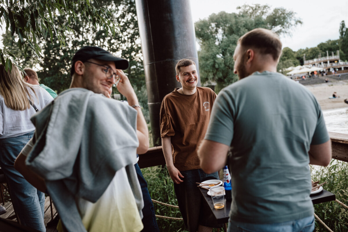 Three men are standing outside by a wooden railing, casually talking and smiling. They are surrounded by greenery, and in the background you can see a sandy area and people near the water. Nearby is a table with drinks and food.  