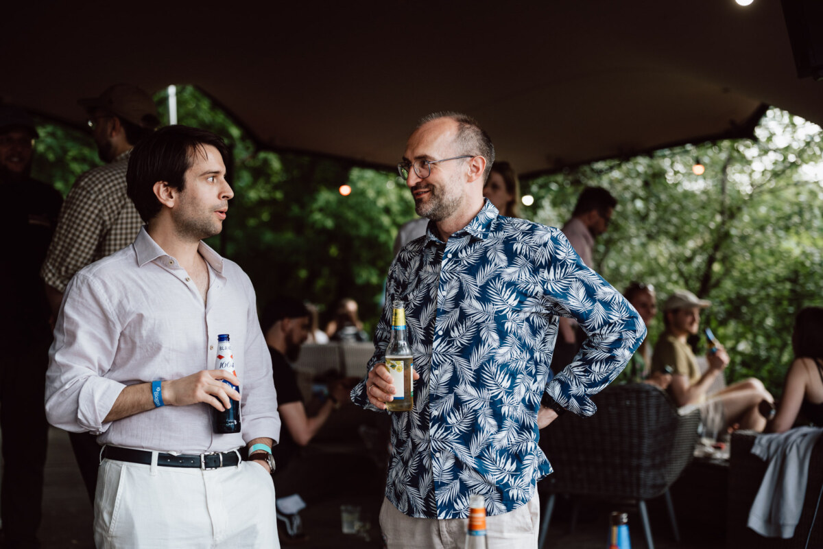 Two men are talking at an outdoor meeting. One is holding a beer, wearing a light-colored shirt, and the other, wearing a blue patterned shirt and glasses, is holding a drink. People are sitting and talking in the background.  
