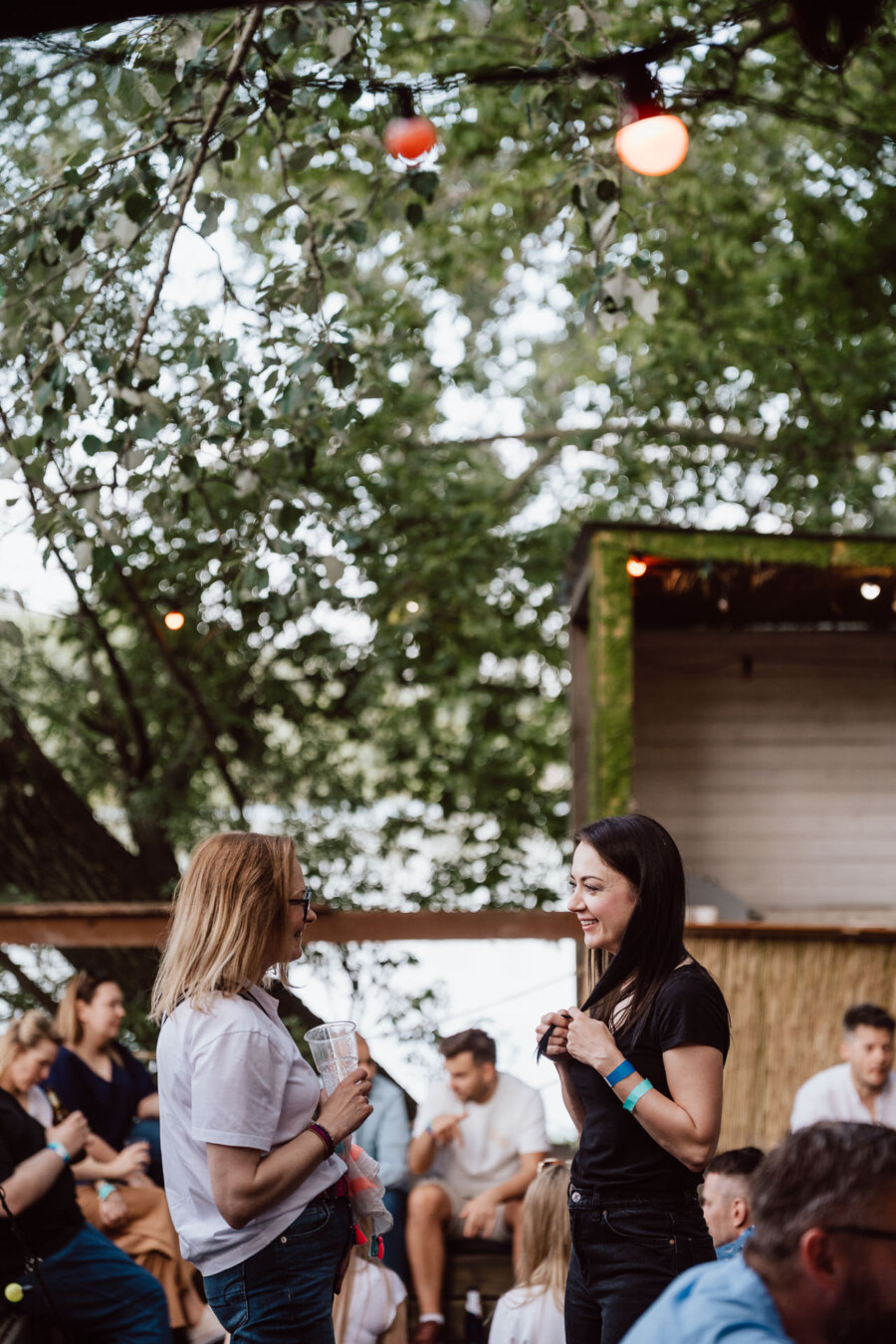 Two women stand and talk at an outdoor meeting under the trees, while groups of people sit and talk in the background. Warm lights hang above them, creating a relaxing, festive atmosphere. 