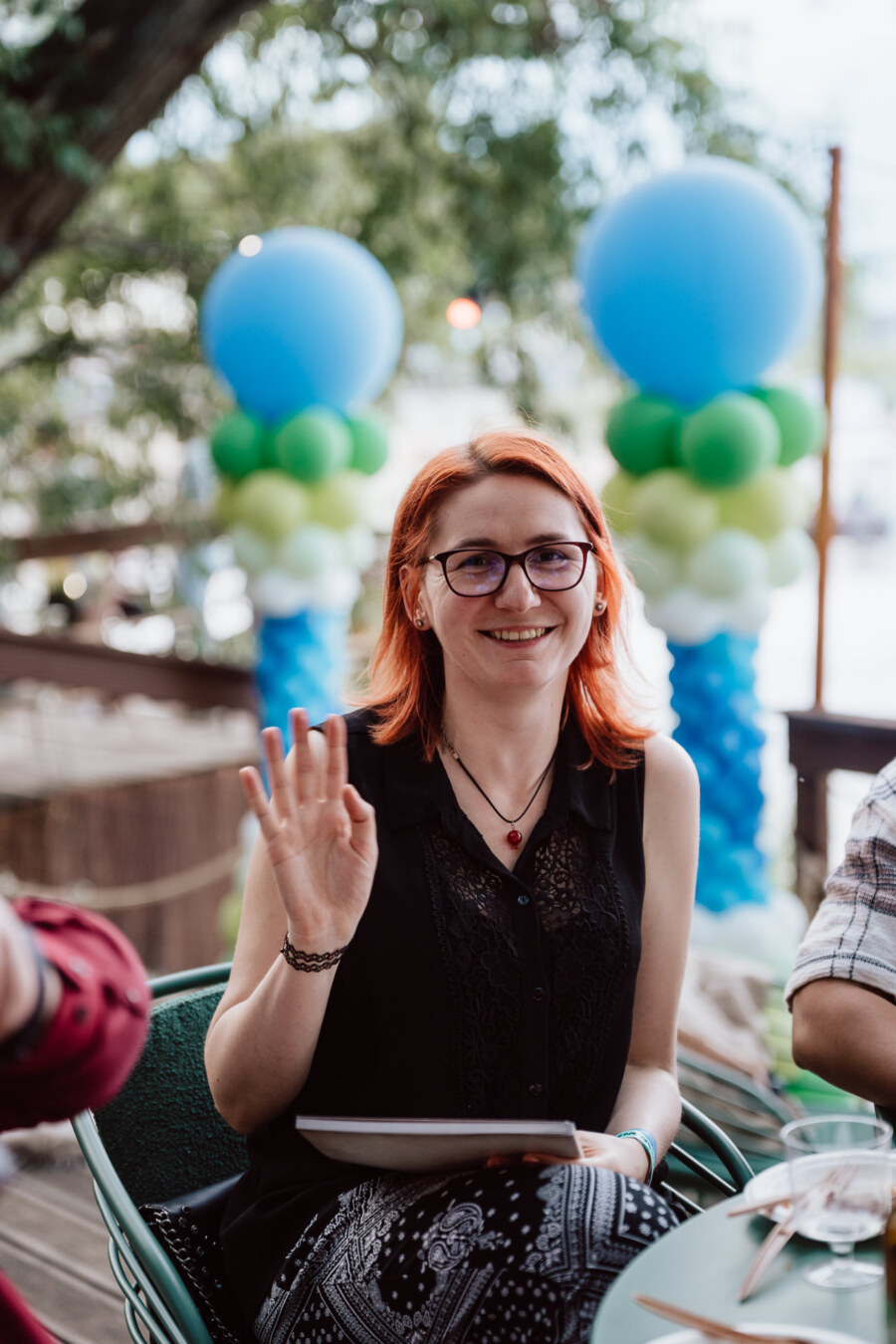 A young woman with red hair and glasses smiles and waves to the camera, sitting outside. Blue and green balloon decorations are visible in the background. 