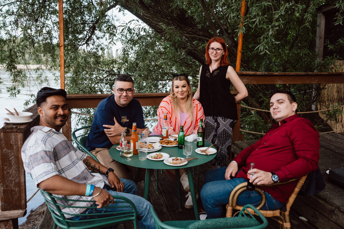 Five people sit and stand around a round green table outside, sharing drinks and food. There are plates of snacks and drinks on the table. Trees and water are visible in the background. Everyone seems relaxed and smiling.   