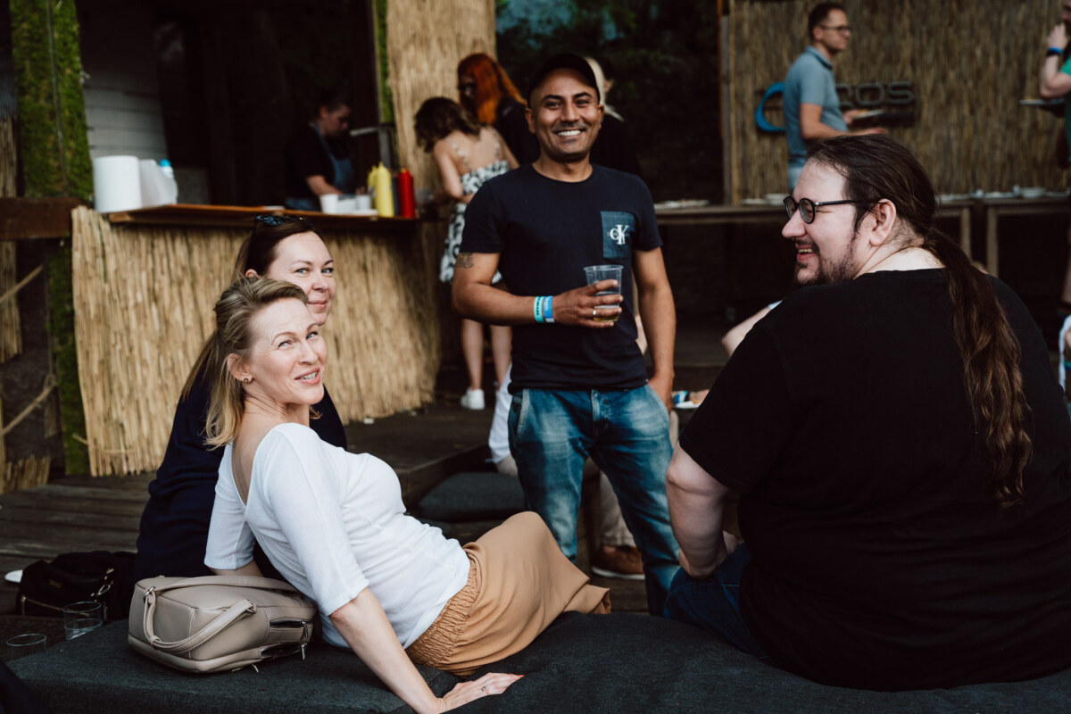 A group of four adults, three seated and one standing, smiling and socializing outside near a wooden structure with drinks, in a casual, relaxed environment. Other people are visible in the background. 