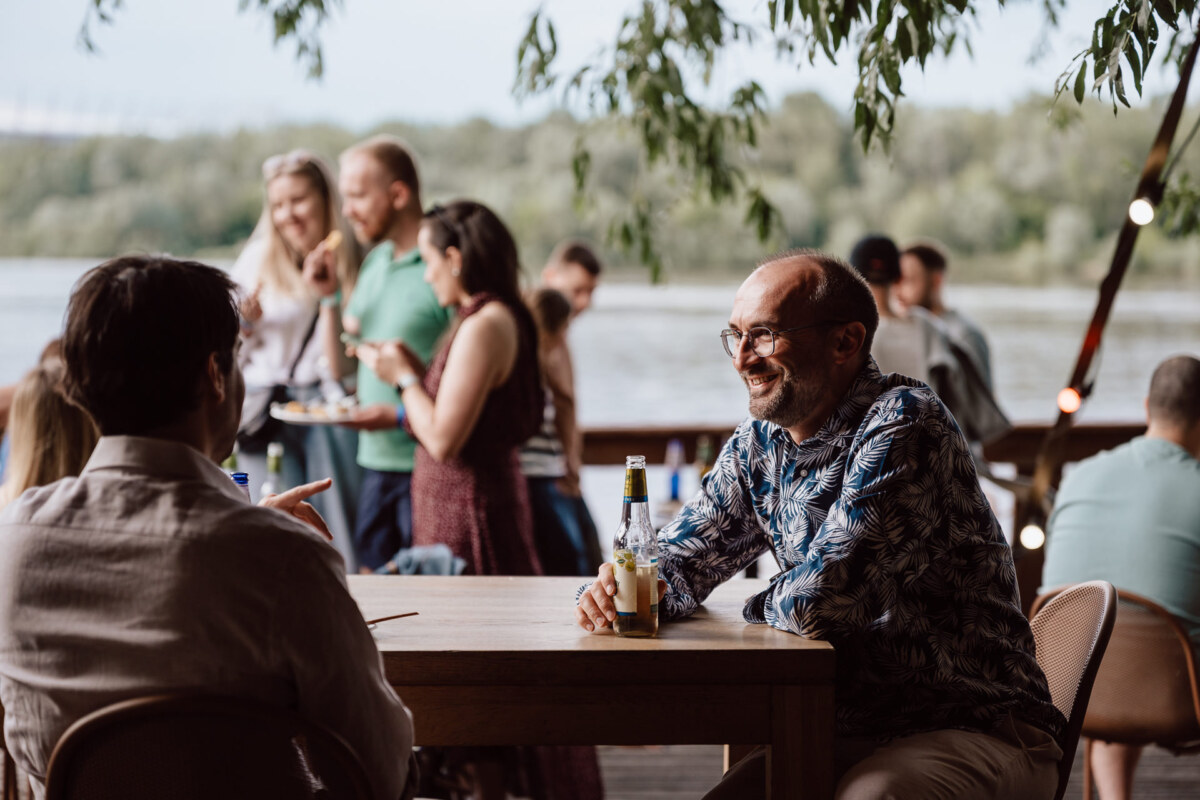 A group of people meet outdoors by a river. In the foreground two men are sitting at a wooden table with drinks, one is smiling. Other people stand and talk in the background, surrounded by greenery.  