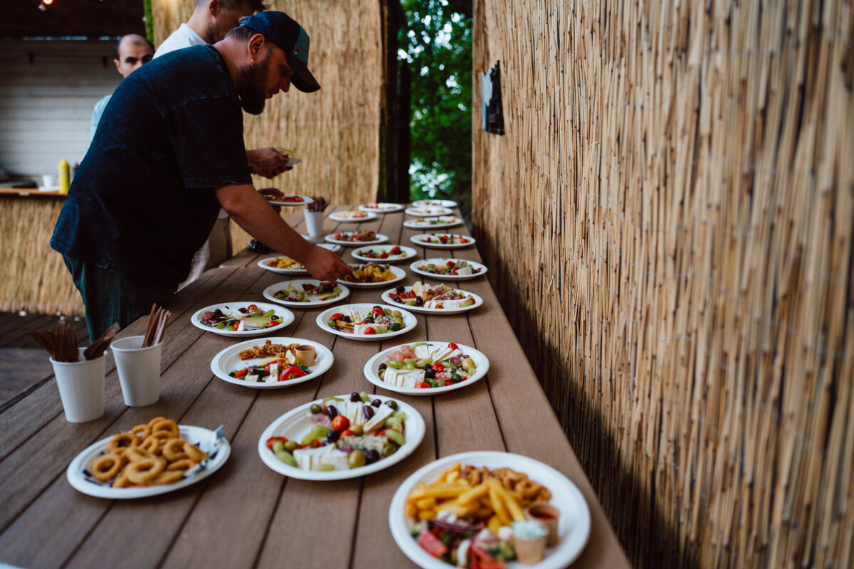 A man arranges plates of assorted food, including French fries, salad and onion rings, on a wooden table outside next to a bamboo wall, while others stand nearby.