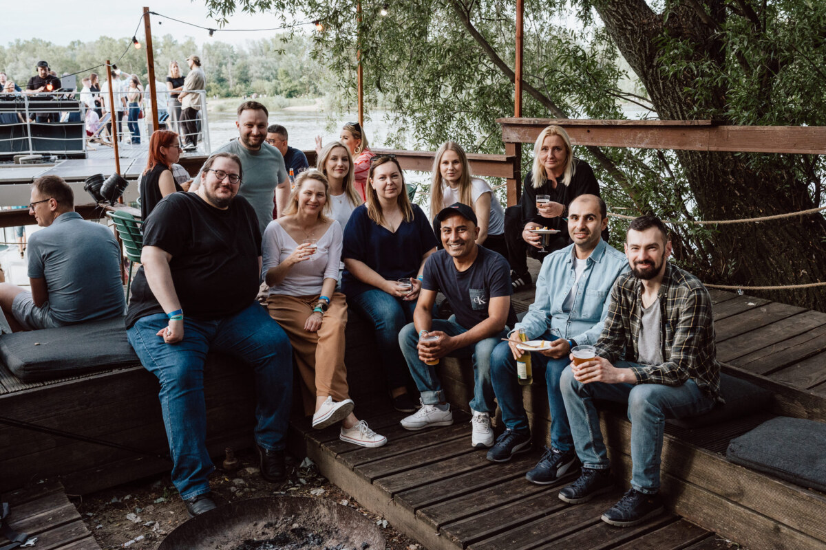 A group of eleven people sit and stand together on a wooden platform by the river, smiling and holding drinks. Trees and other people are visible in the background, creating a relaxing outdoor atmosphere. 
